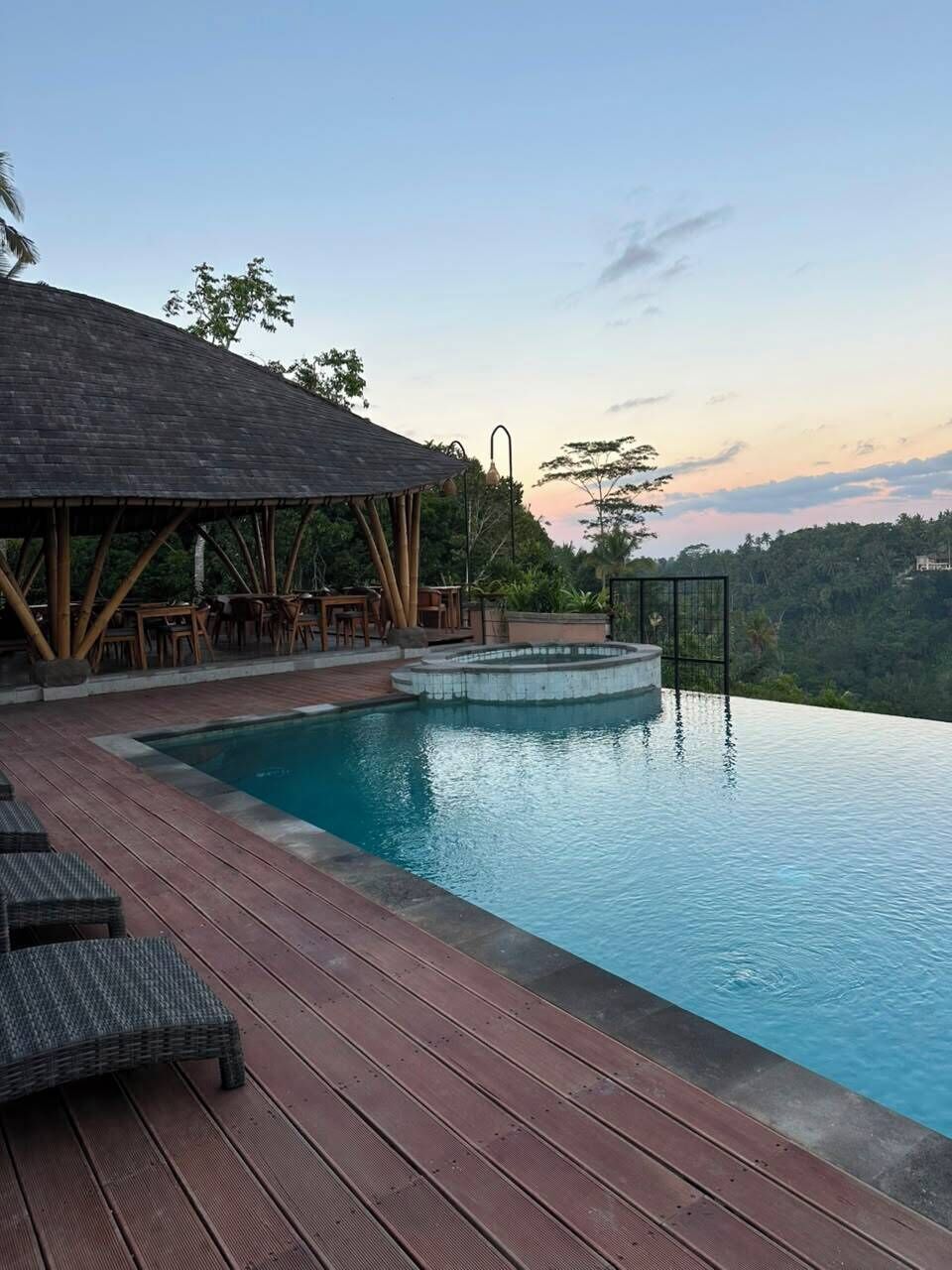 Una piscina infinita con un deck de madera y sillas de descanso, rodeada de exuberante vegetación y un cielo despejado.