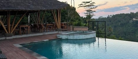 An infinity pool with a wooden deck and lounge chairs, surrounded by lush greenery and a clear sky.
