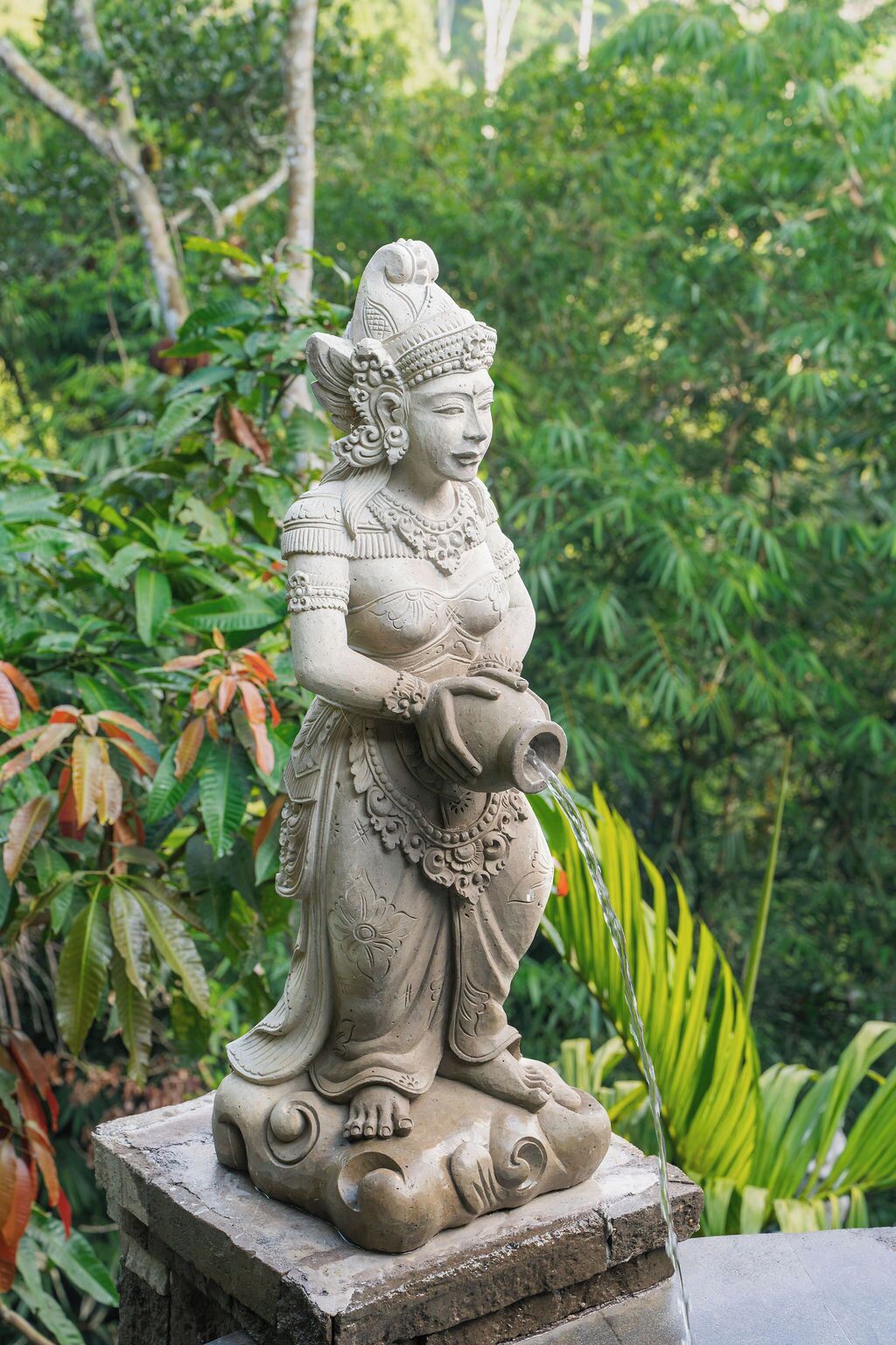 A stone statue of a figure seated on a pedestal with a water spout, surrounded by lush greenery.
