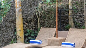 Dos sillones de mimbre con toallas, un paraguas decorativo y una piscina con un muro de piedra y follaje al fondo.