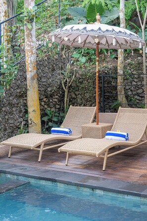 Two wicker lounge chairs with towels, a decorative umbrella, and a poolside setting with a stone wall and foliage in the background.