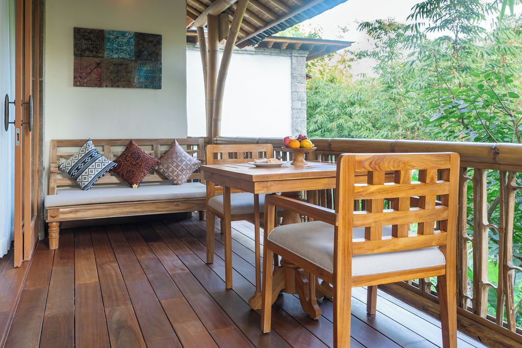 A wooden deck with a bench and chairs, a table with fruit, and a view of greenery.