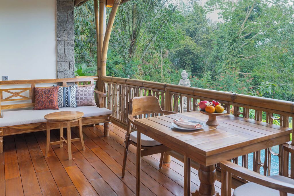 Une terrasse en bois avec un espace salon, une table garnie de fruits et une statue en arrière-plan.