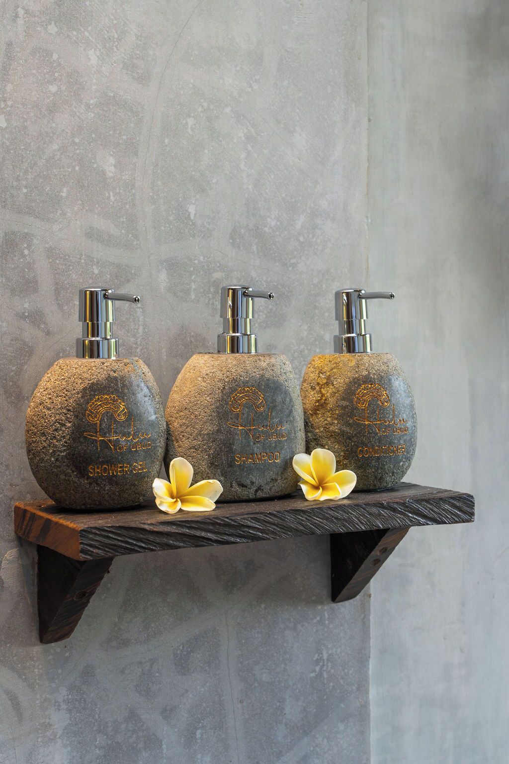 Three stone bottles with pumps, labeled for shower gel, shampoo, and conditioner, on a wooden shelf against a textured wall.