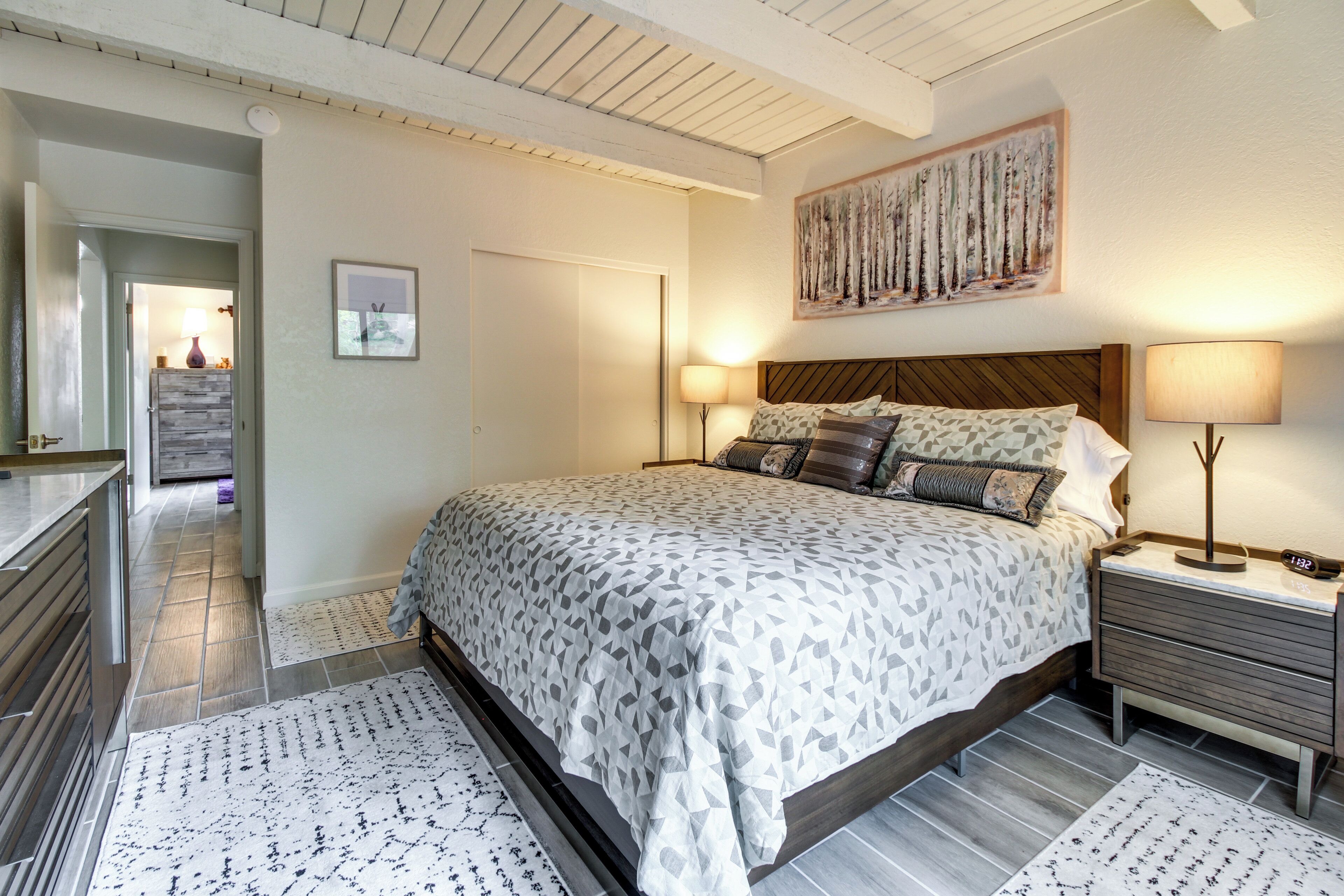 A bedroom with a bed, nightstands, a painting, and a door leading to another room.
