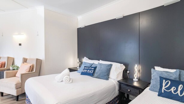 A hotel room with a large bed, two bedside tables, a chair, and a wall-mounted light fixture. - Room-deluxe Family Room - Sleeps 5 (Sydney)