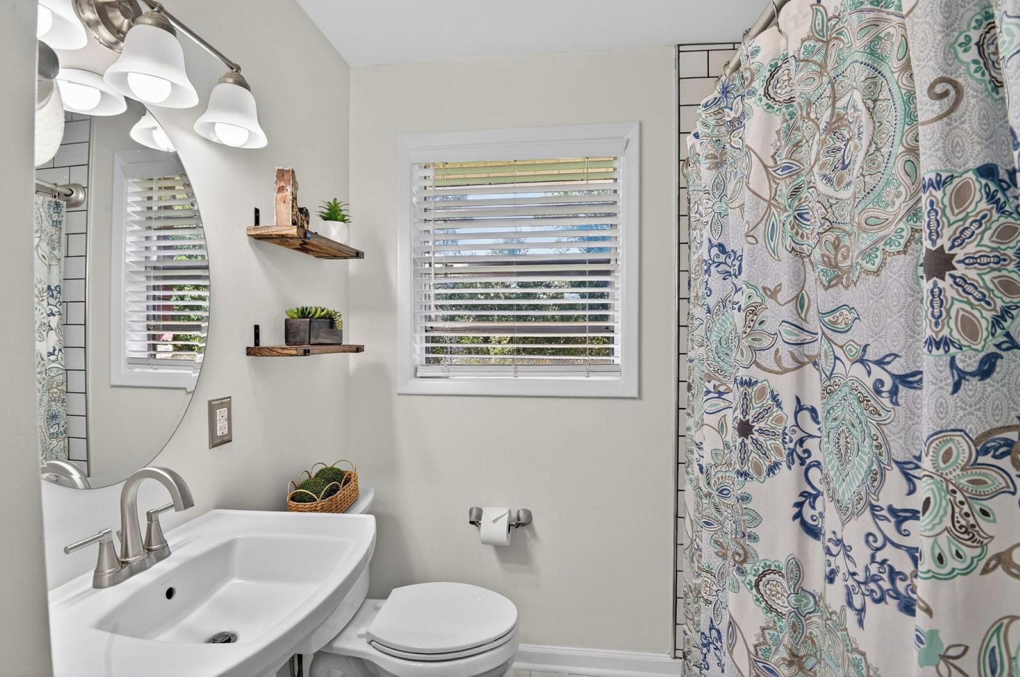 A bathroom with a sink, mirror, window with blinds, toilet, and a shower curtain.