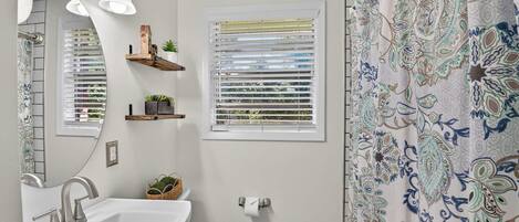 A bathroom with a sink, mirror, window with blinds, toilet, and a shower curtain.