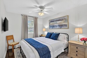 Ein Schlafzimmer mit einem Bett, einem Sessel, einem Nachttisch mit Lampe, einem an der Wand befestigten Fernseher und einem Deckenventilator.