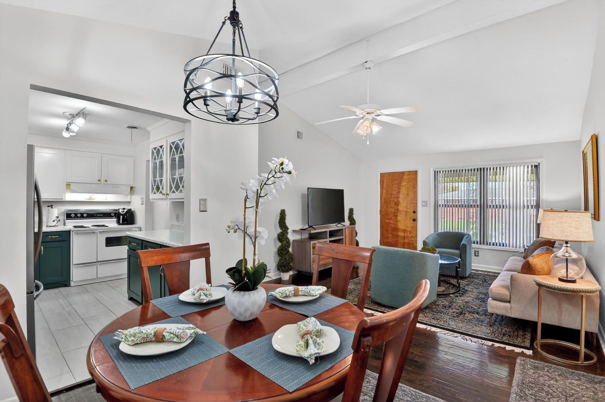 A modern kitchen with white cabinets, a dining area with a wooden table and chairs, a living room with a sofa and a TV, and a ceiling fan.