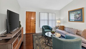 A living room with a television, a wooden door, a window with blinds, a sofa, a chair, a small table, and a lamp. - Golf & Chill! Family-Friendly Augusta Home (Augusta)