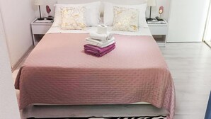Een opgemaakt bed met een roze deken, twee witte kussens en een stapel handdoeken.