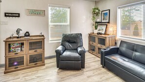 A living room with a leather sofa, a leather armchair, and a wooden cabinet with glass doors. There are two windows with blinds, a welcome sign, and a framed picture on the wall. - Room for 6 Cars on Balloon Fiesta Weekend (Albuquerque)