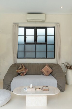 Ein modernes Wohnzimmer mit großem Fenster, einer Couch mit Kissen, einem Couchtisch mit Dekoration und einer Topfpflanze.