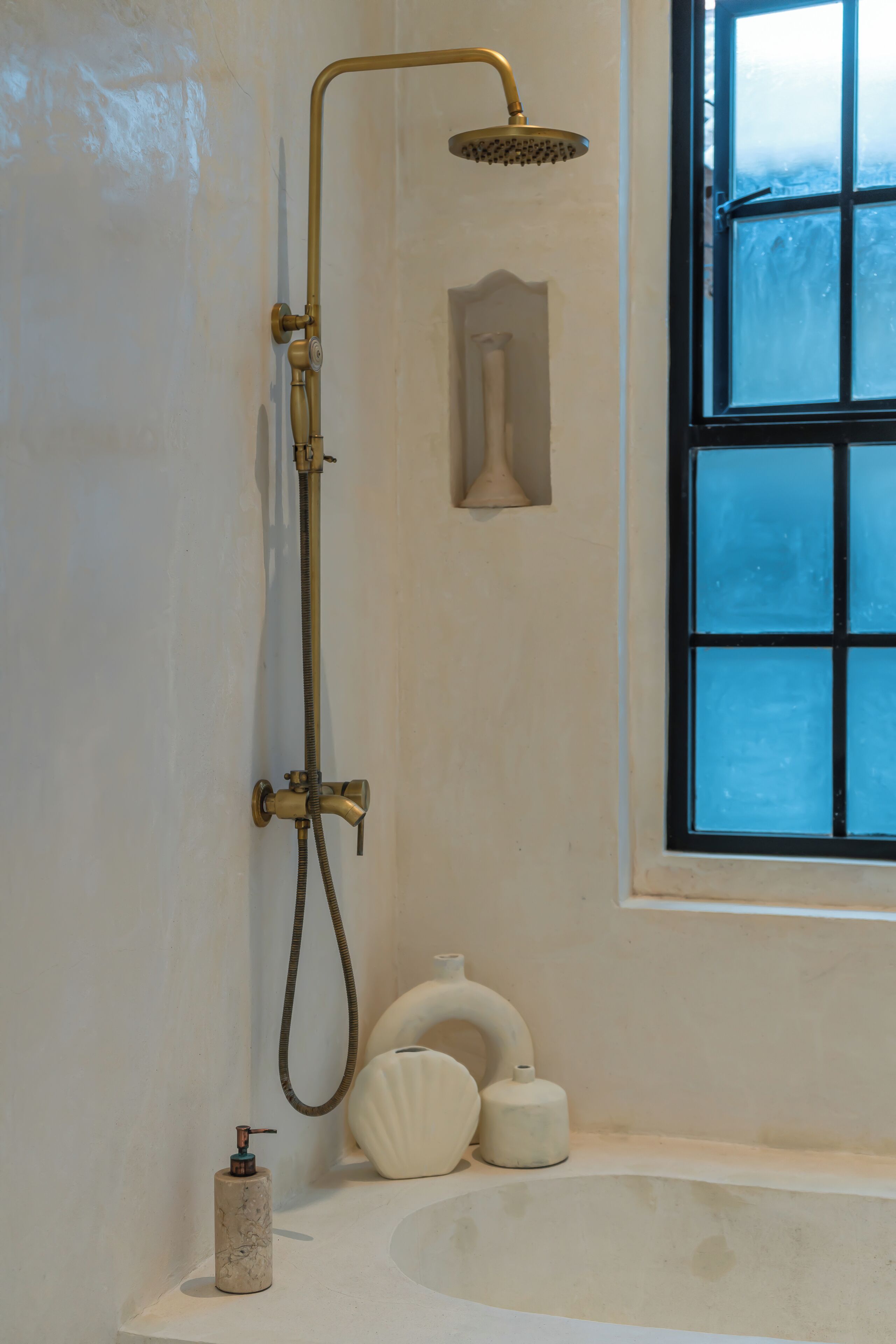 A bathroom with a bathtub, a shower with a gold-colored showerhead, a window with frosted glass, and a soap dispenser.