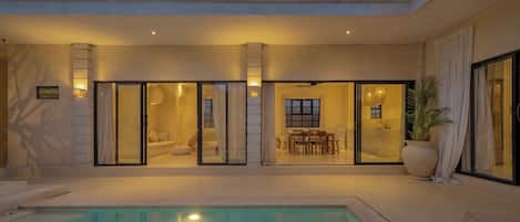 A modern outdoor pool area with a glass enclosure, a dining area, and a large potted plant.