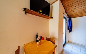 A room with a wooden ceiling, a television mounted on the wall, a table with a yellow tablecloth, a bottle of wine, and a vase with flowers. - Amazing apartment in Viganj with WiFi (Viganj)