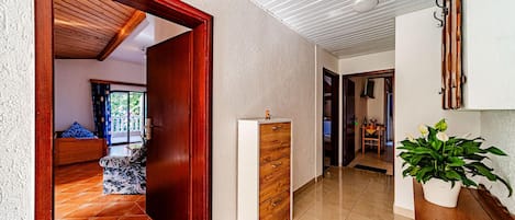 A hallway with a wooden door leading to a room with a bed and a view of greenery outside. There is a white cabinet with drawers and a potted plant on a wooden table.