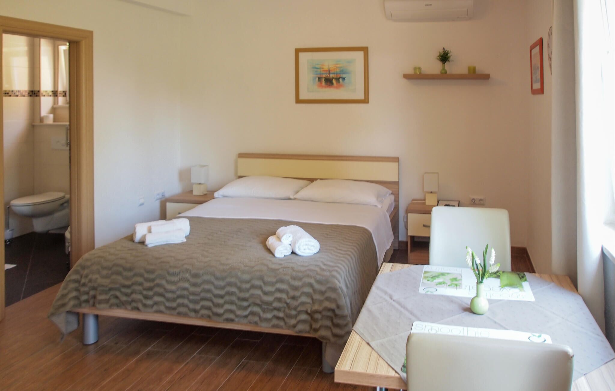 A hotel room with a bed, bedside tables, a chair, a small table with a vase, a painting on the wall, and a bathroom visible through an open door.