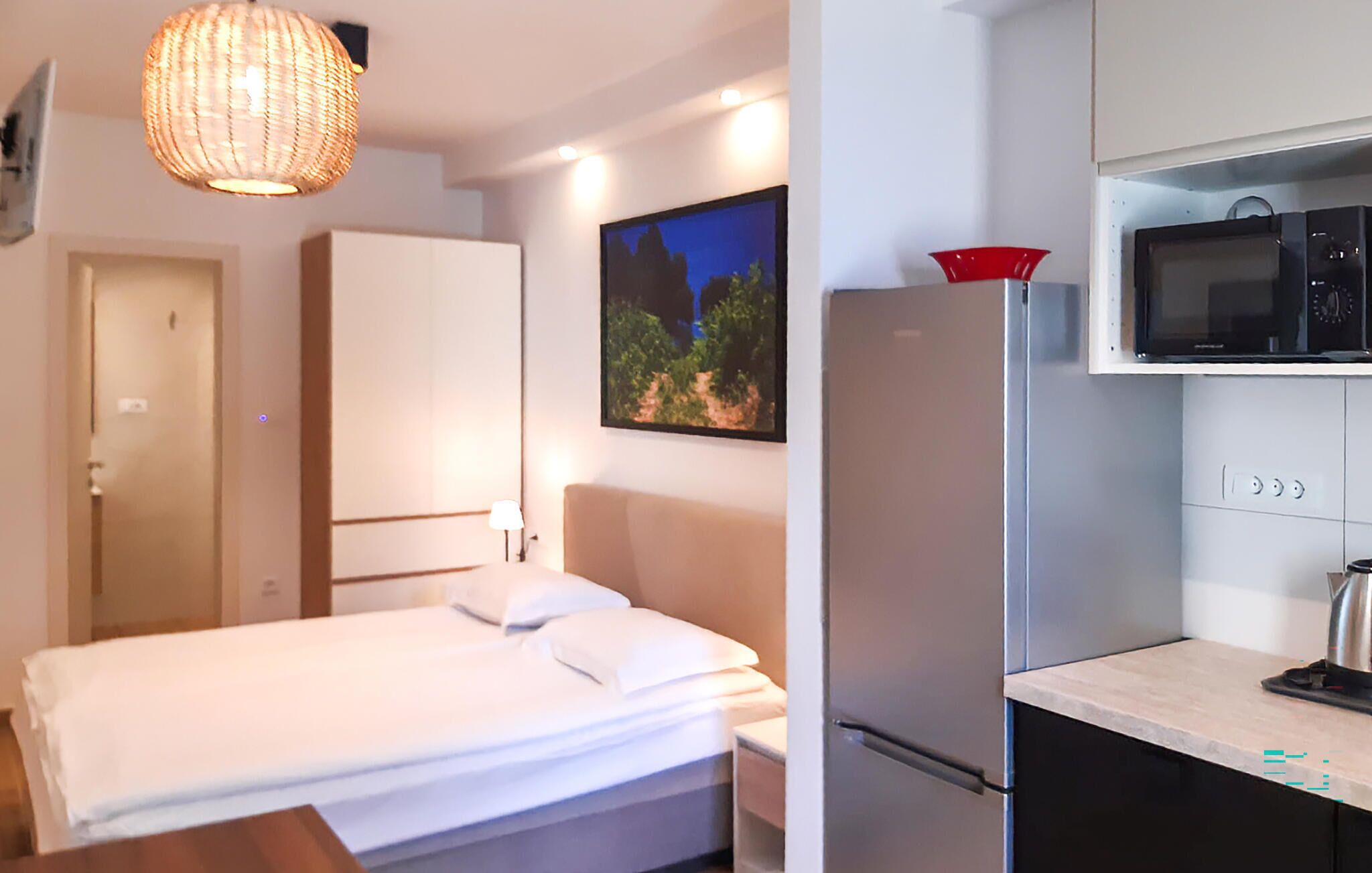 A hotel room with a bed, a wardrobe, a painting, a refrigerator, and a kitchenette.