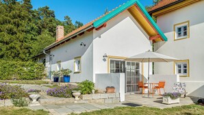 Ein Haus mit geneigtem Dach, einer Terrasse mit Gartenmöbeln und einem Garten mit verschiedenen Pflanzen und Blumen.