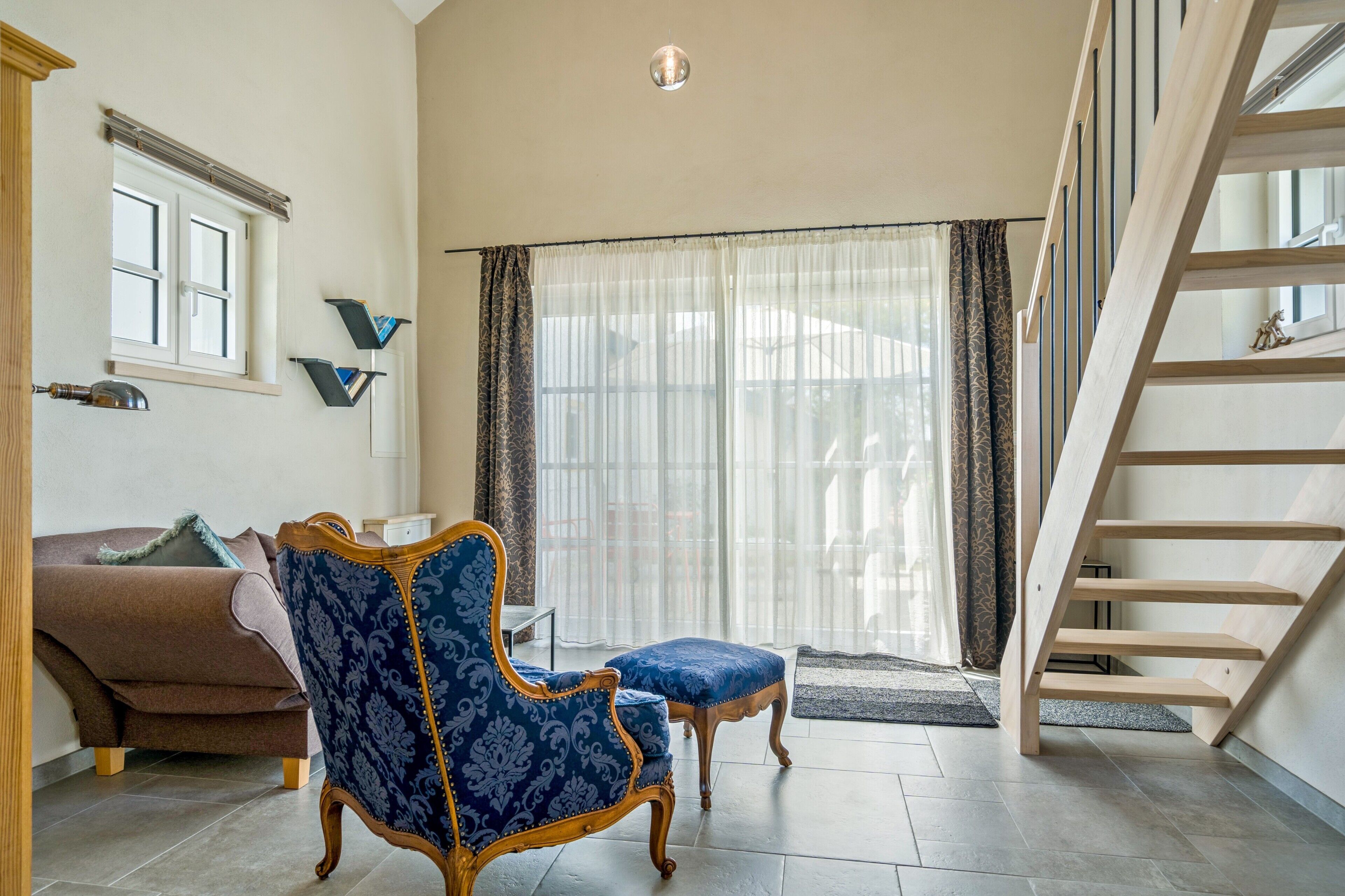 Un salon spacieux avec un fauteuil à motifs bleus, un canapé marron, un escalier en bois et une grande porte-fenêtre coulissante donnant sur un espace extérieur.