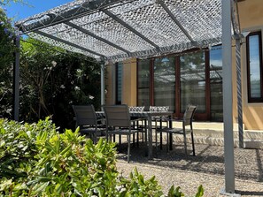 Eine sonnige Terrasse mit Tisch und Stühlen, umgeben von Pflanzen und einer Pergola.