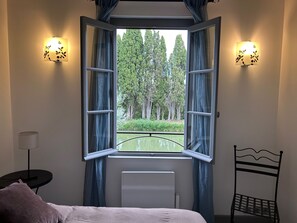 Een slaapkamer met een open raam, een bed, een stoel, een nachtkastje en wandlampen.