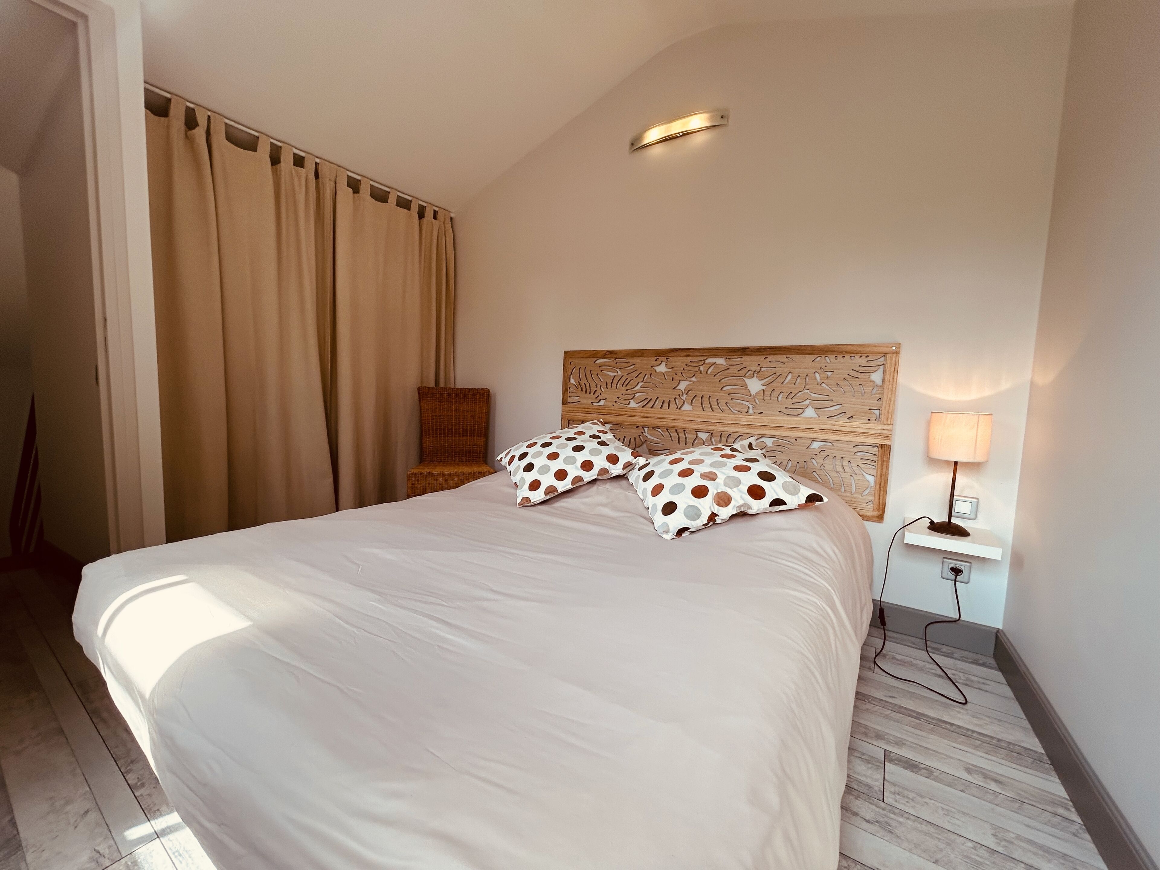 Habitación de hotel con una cama grande, cabecera de madera, mesita de noche con lámpara y una silla.