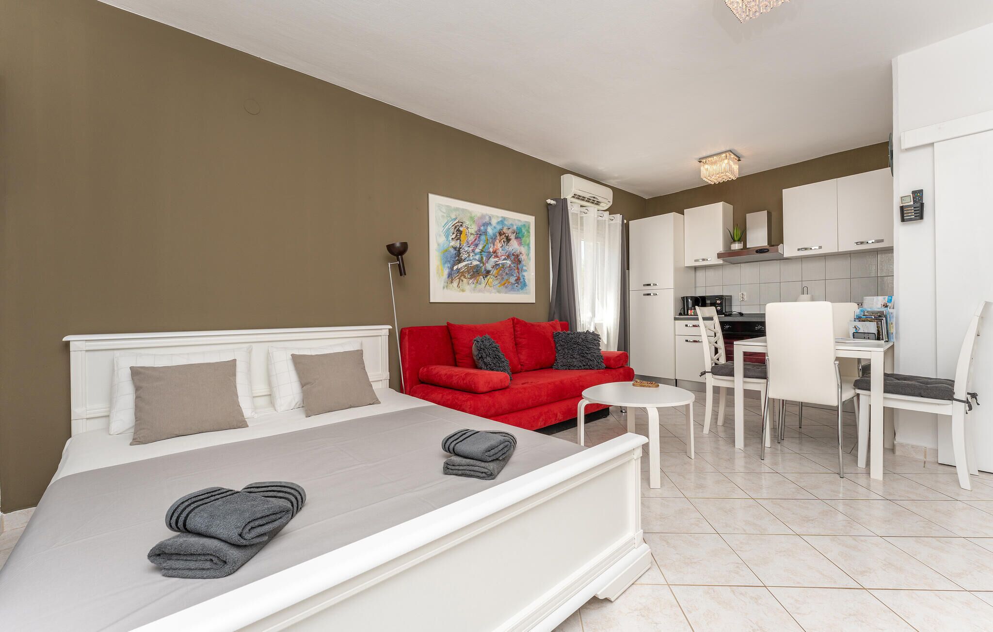 Un dormitorio moderno con una cama, un sofá rojo, una mesita y una kitchenette al fondo.