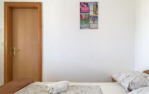 Ein Schlafzimmer mit Holztür, einem Bett mit weißer Matratze und Kissen sowie einer Wand mit einem farbenfrohen Plakat.