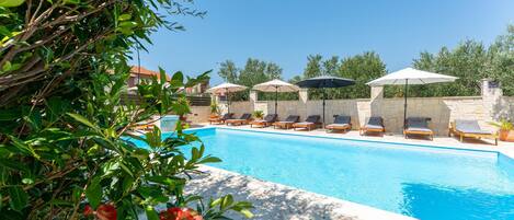 A poolside area with lounge chairs and umbrellas, surrounded by greenery and a stone wall.