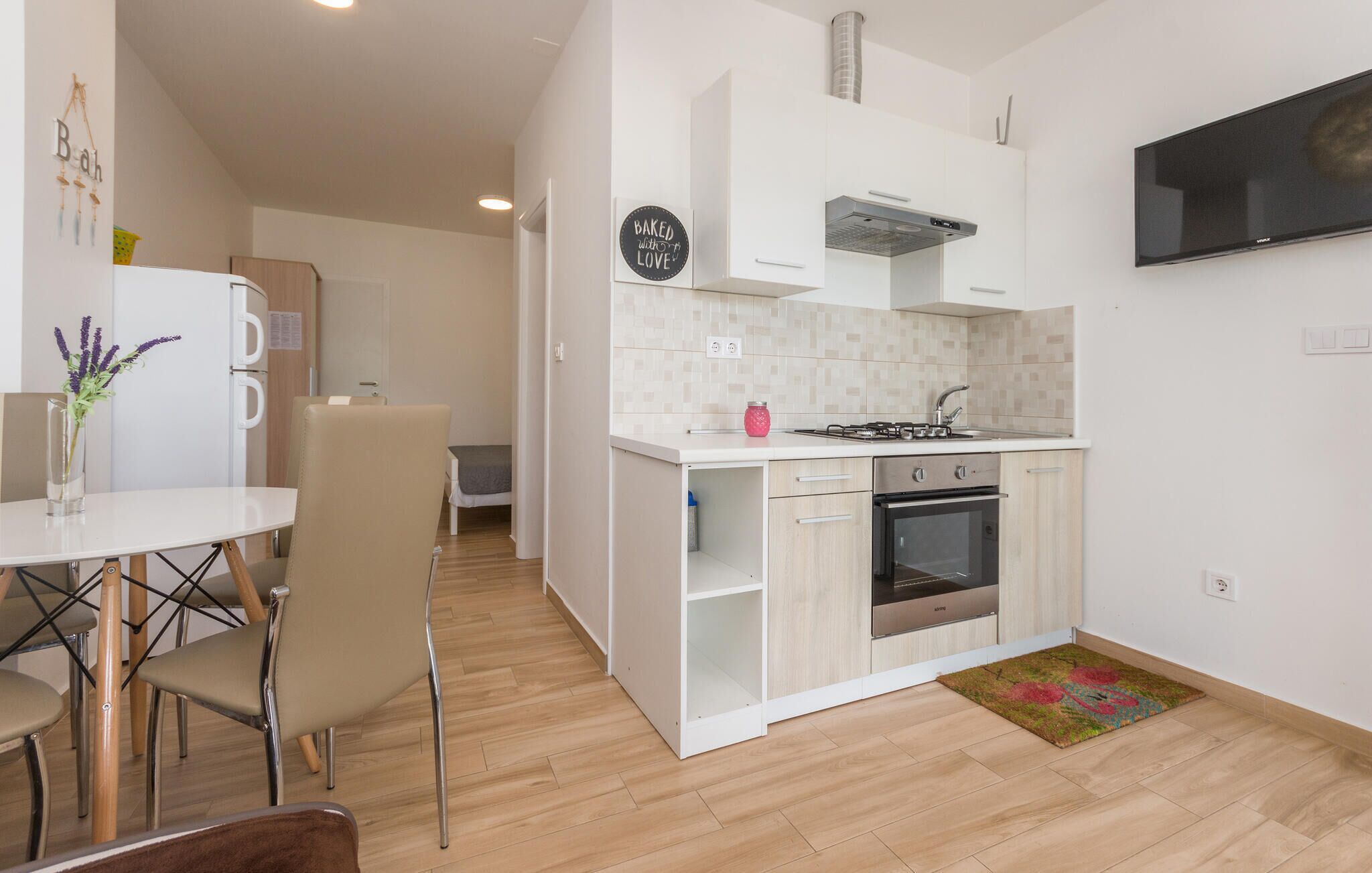 Une cuisine compacte dotée d’un four encastré, un coin repas avec une table et des chaises, et un petit téléviseur fixé au mur.