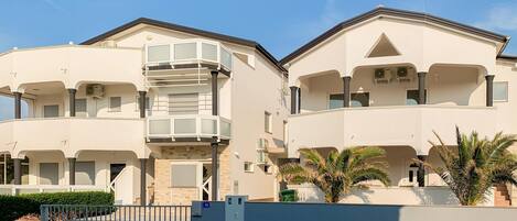 A two-story residential building with a balcony on the first floor, a gated entrance, and a blue wall with a number sign.