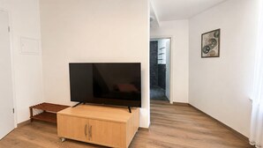 A modern living room with a flat-screen TV on a wooden cabinet, a small seating area, and a hallway leading to another room. - Zum Rheinberg – Komfort Studio (Braubach)