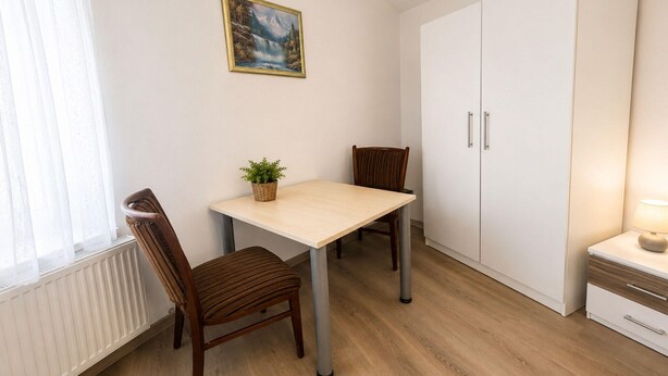 A small room with a dining table, two chairs, a white wardrobe, a radiator, and a window with sheer curtains. - Zum Rheinberg – Komfort Studio (Braubach)