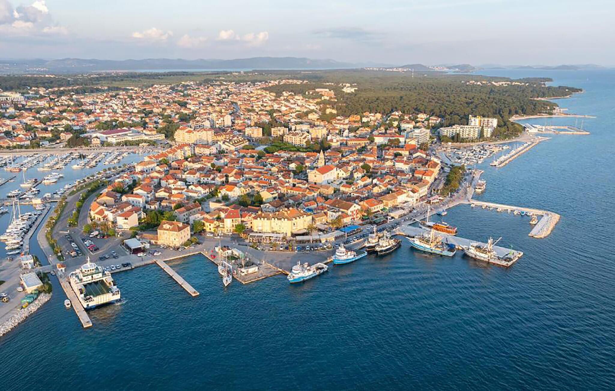 An aerial view of a coastal town with a marina, dense housing, and a clear view of the sea.