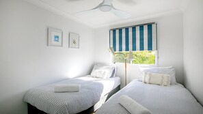 A bedroom with two single beds, a window with a striped blind, and a ceiling fan. - Sunny Bayside Cottage - Walk to bay and village (HARDYS BAY)