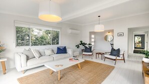 Ett modernt vardagsrum med en vit soffa, ett soffbord och en matta. Det finns ett stort fönster med utsikt över träd och en dörr som leder till ett annat rum.