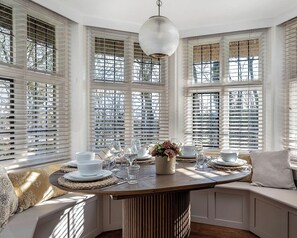 Un coin repas avec une table ronde dressée pour quatre personnes, entouré de meubles blancs et de grandes fenêtres équipées de stores.
