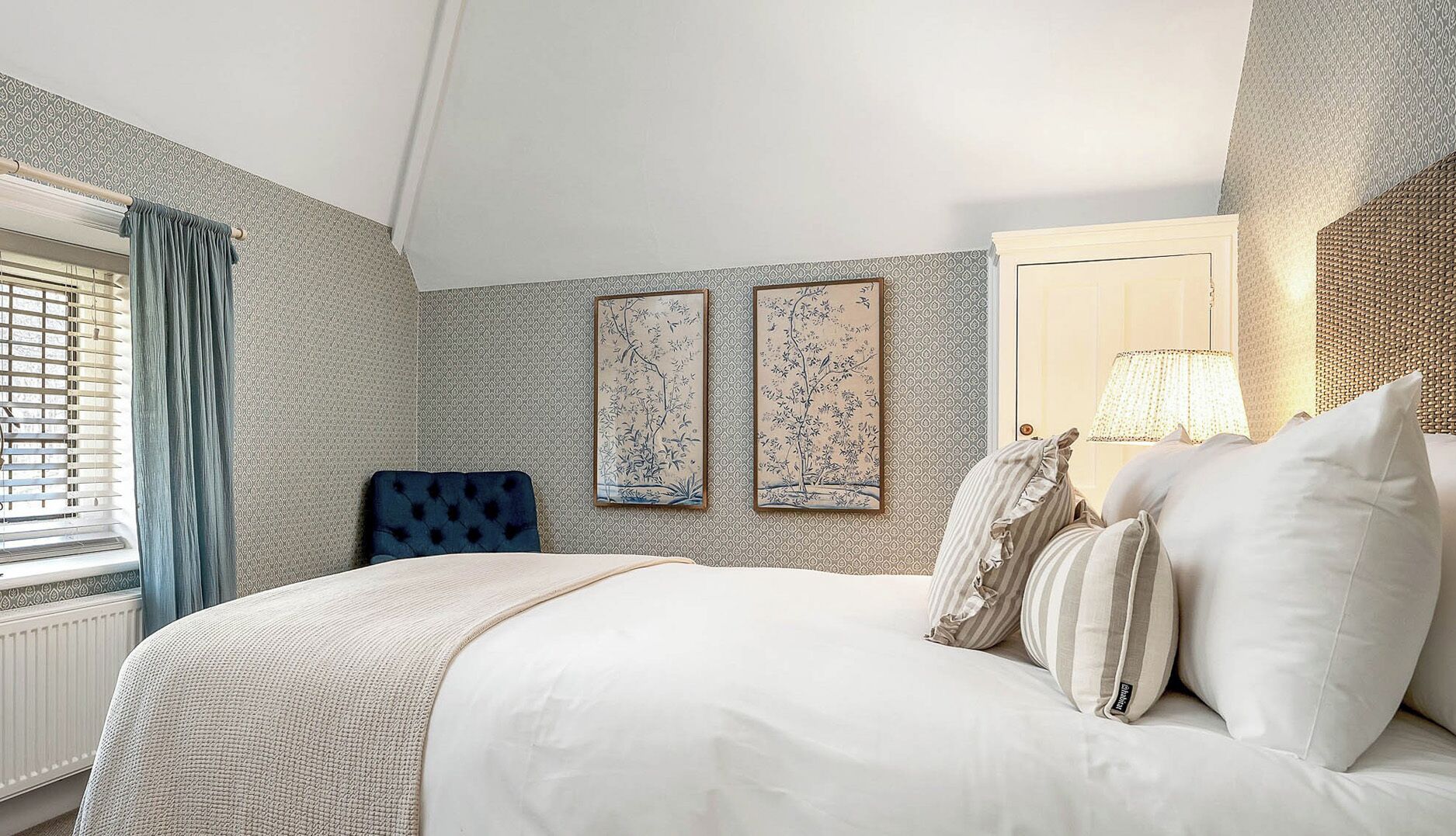 A hotel room with a large bed, a window with blinds, a blue chair, and two framed artworks on the wall.