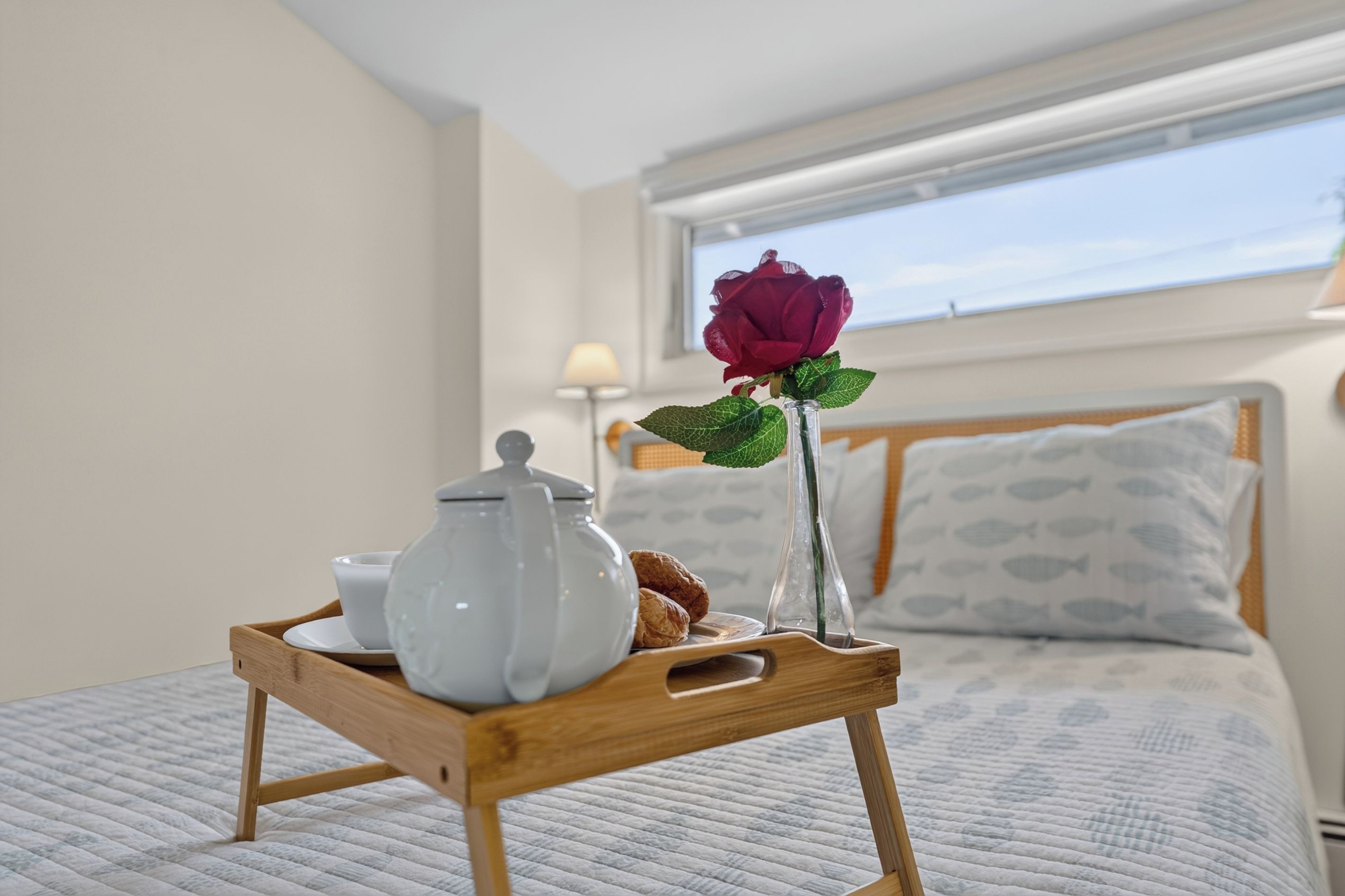 A wooden tray with a teapot, a cup, and a rose on a bed with a fish-patterned pillow and a window with a view.