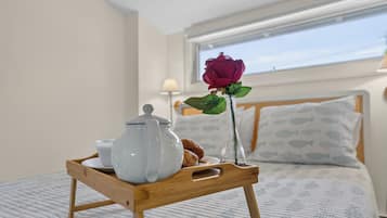 A wooden tray with a teapot, a cup, and a rose on a bed with a fish-patterned pillow and a window with a view.