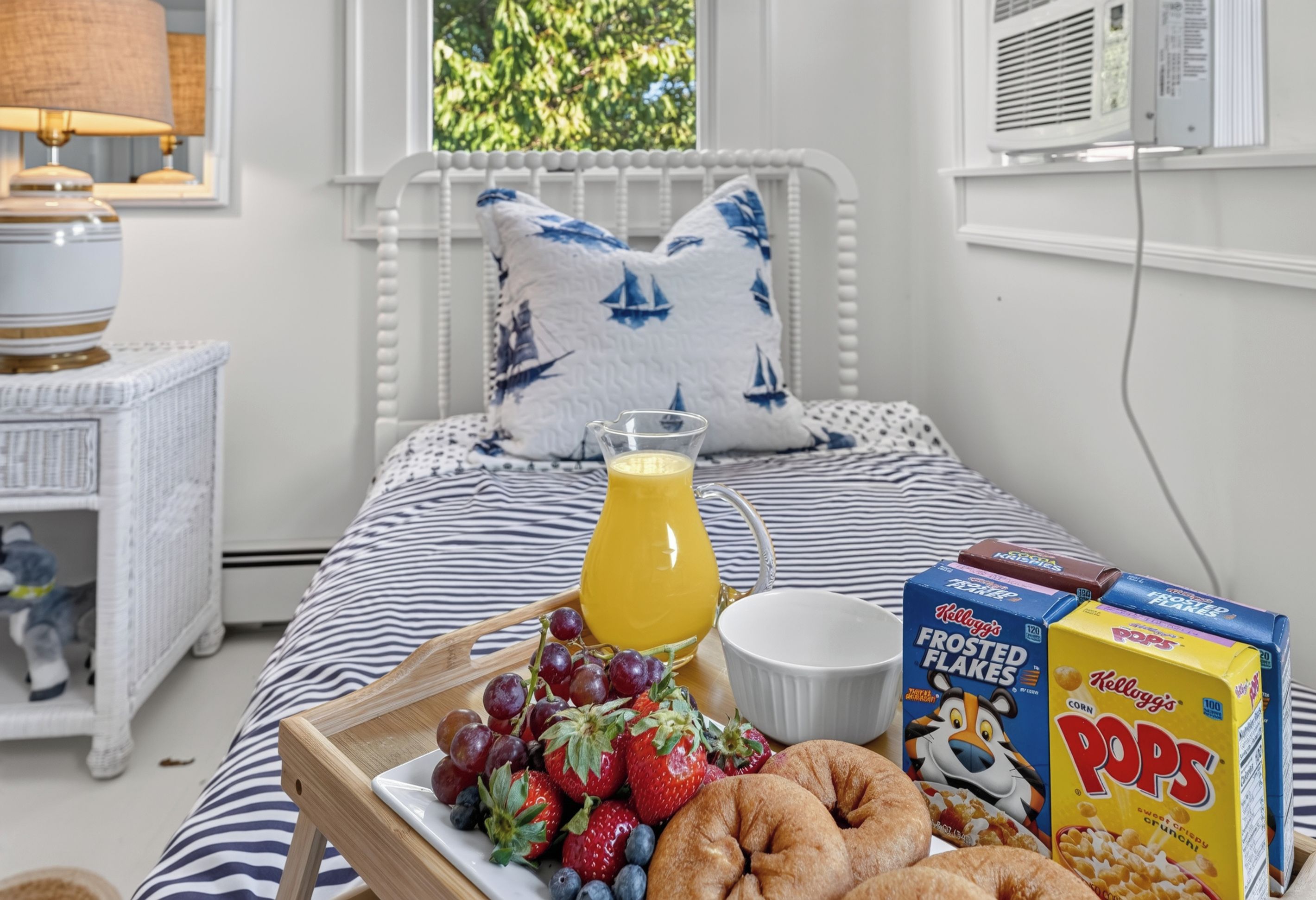 A neatly made bed with a striped bedspread, a bedside table with a lamp, a tray with fruits and a glass of orange juice, and cereal boxes on the bed.