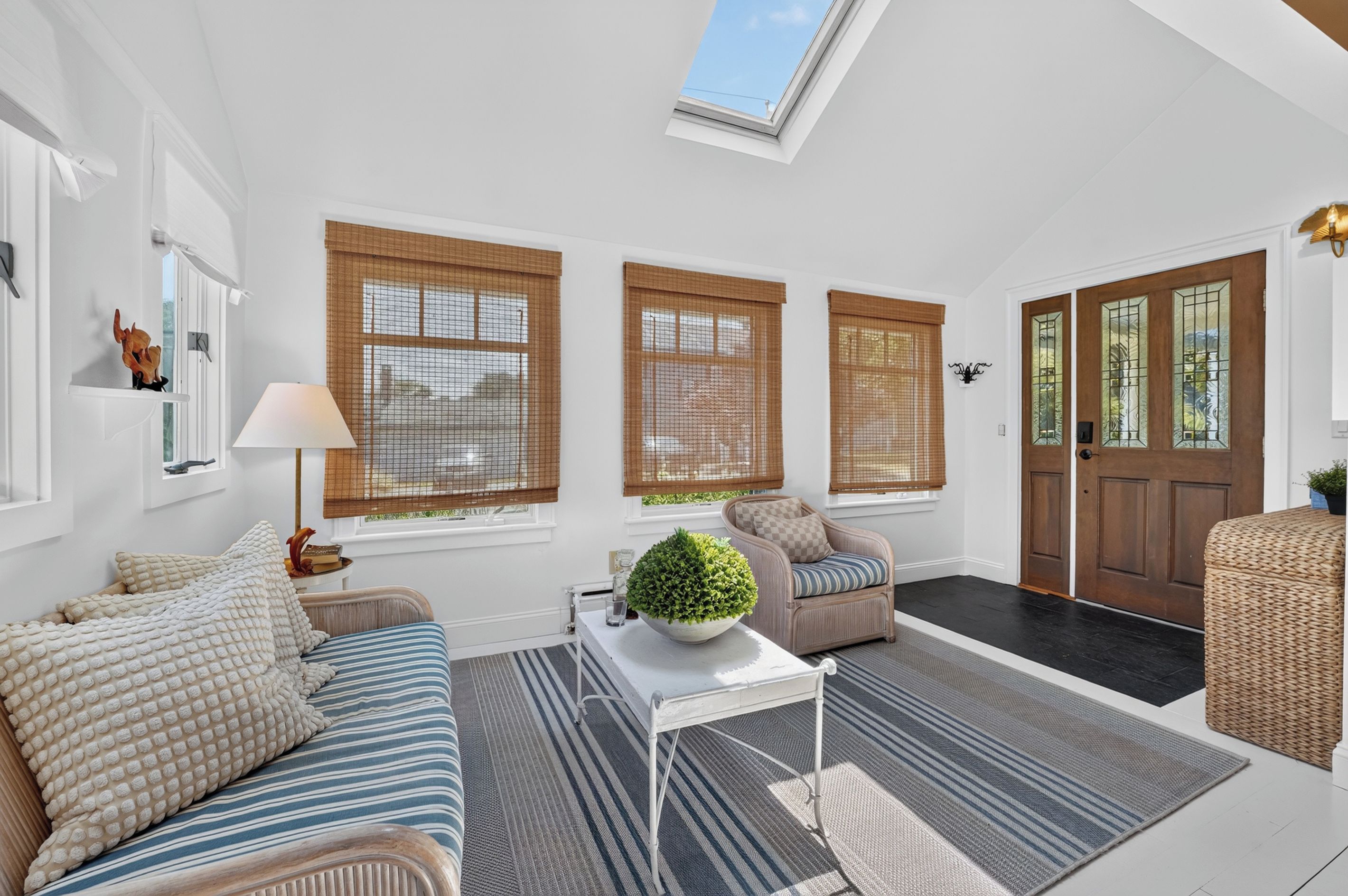 A well-lit living room with a sofa, armchair, and a small table with a plant. There are large windows with blinds, a door with glass panels, and a skylight.