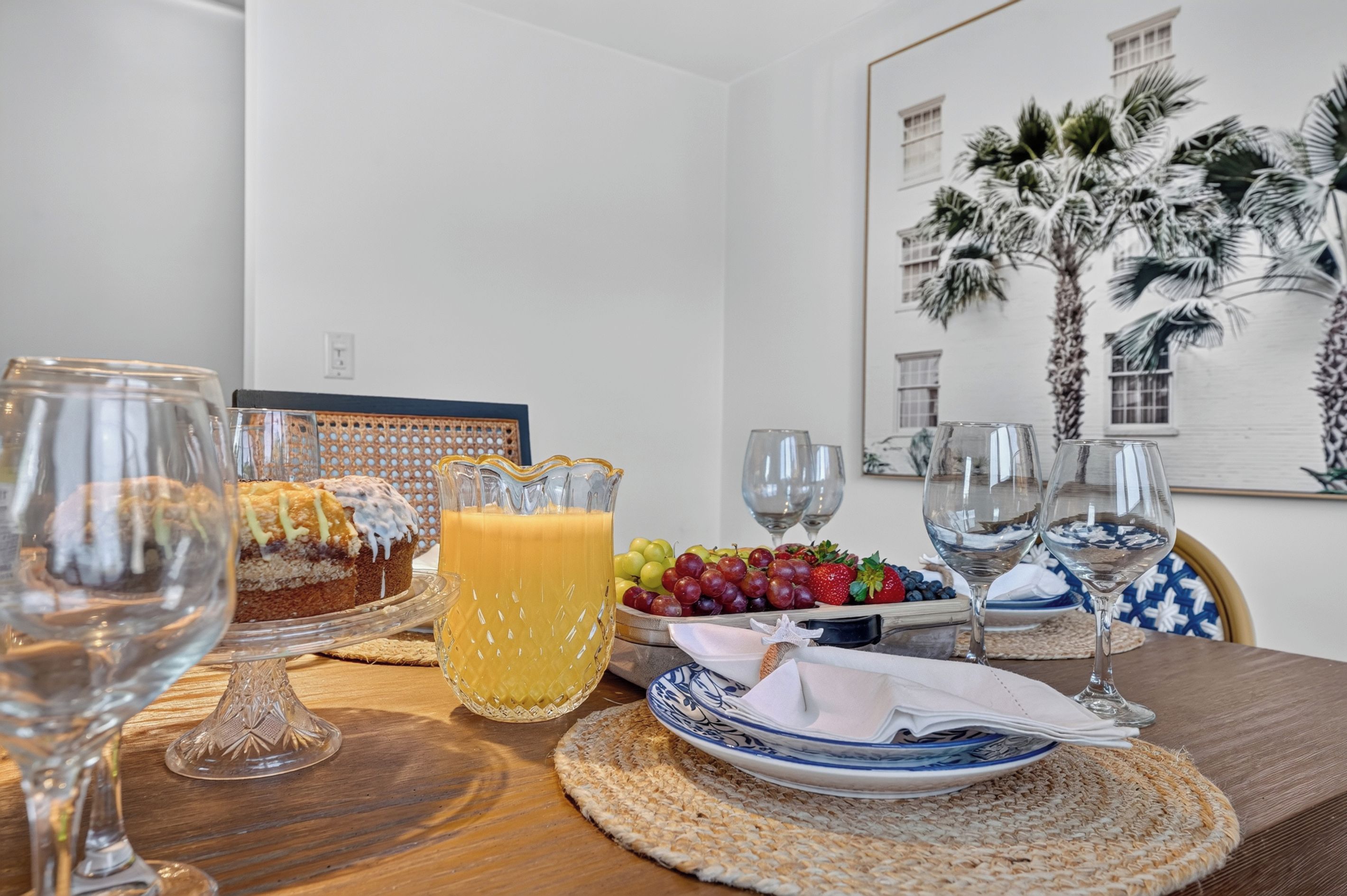 Une table à manger avec un plateau de fruits, une carafe de jus d’orange et des verres à vin.