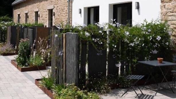 Un edificio con tetto spiovente, facciata in mattoni e un vialetto lastricato che conduce a un'area relax con tavolo e sedie.