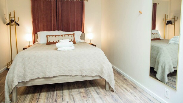 Uma cama bem arrumada com manta e travesseiros brancos, assoalho de madeira, um espelho grande e luminária fixada na parede.