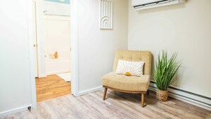 Quarto com porta para banheiro, ar condicionado fixado na parede, cadeira de vime com almofada e planta em vaso.