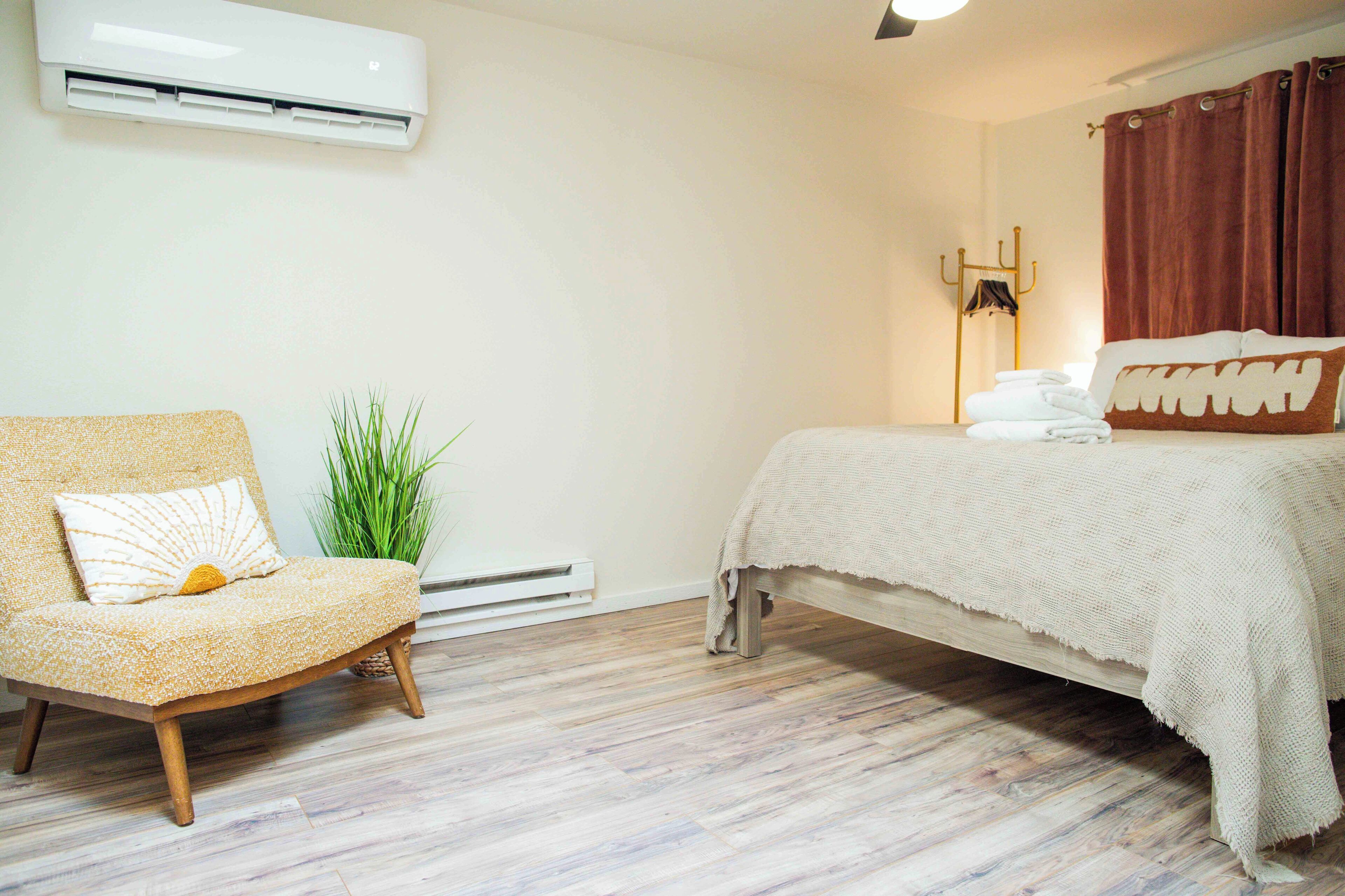 A bedroom with a bed, a chair, a plant, and an air conditioning unit.